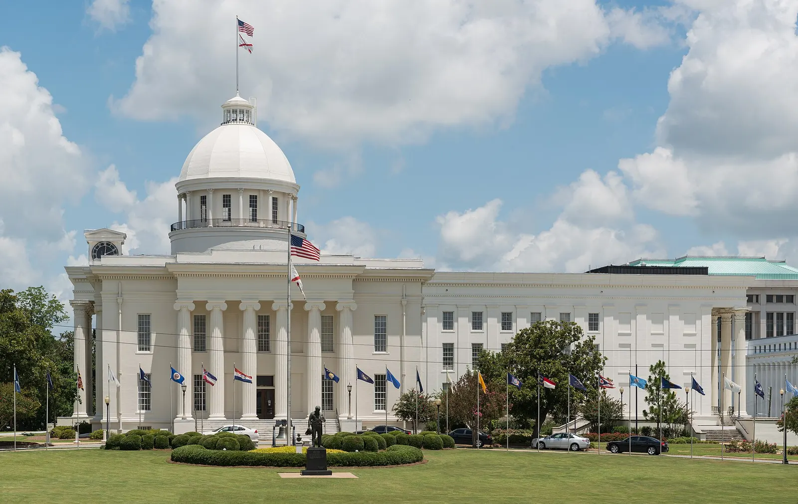 The Alabama State Capitol in Montgomery: a white domed building with a columned front, flagpoles and a bronze statue on the lawn under a partly cloudy sky