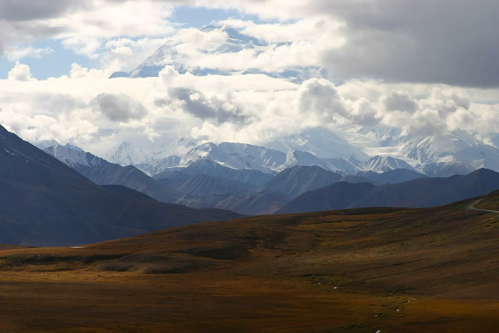 Snow-covered mountains partly hidden by clouds above brown tundra, in Denali National Park and Preserve, Alaska