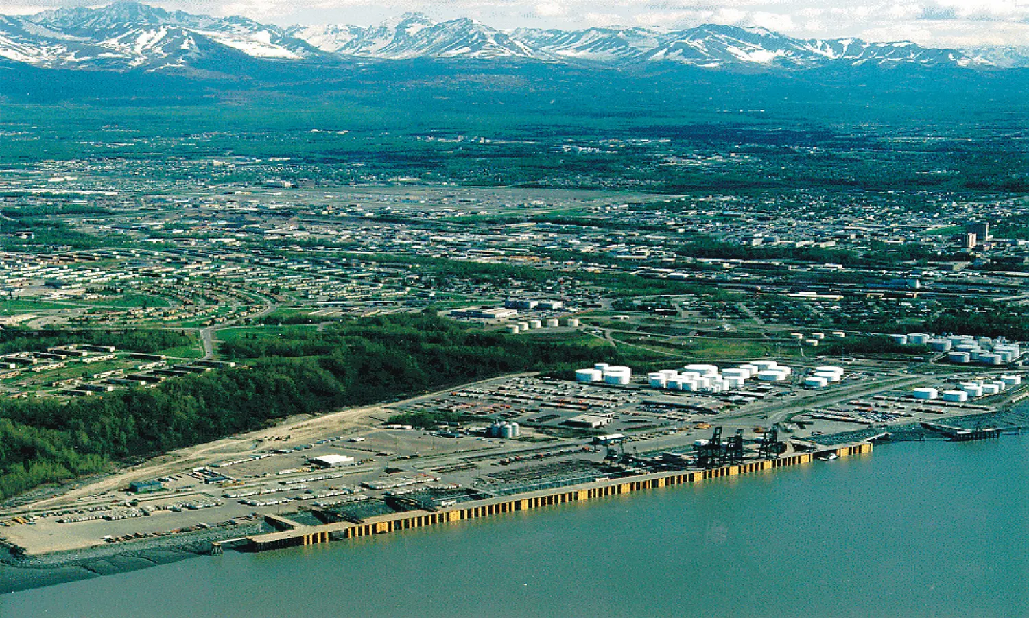 Aerial view of a port waterfront on the Anchorage shoreline with docks, storage tanks and container yards, and snow-capped mountains behind the city