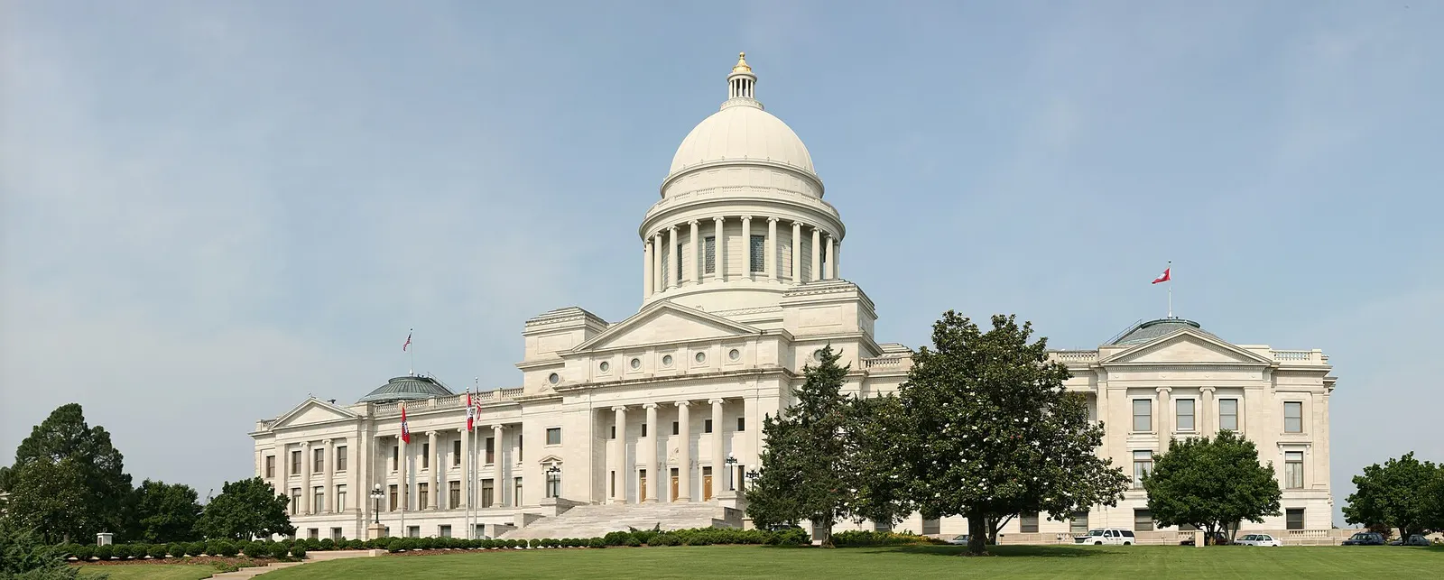 A wide view of a white stone capitol building with a central dome and a columned entrance, seen across a lawn with trees under a pale blue sky.