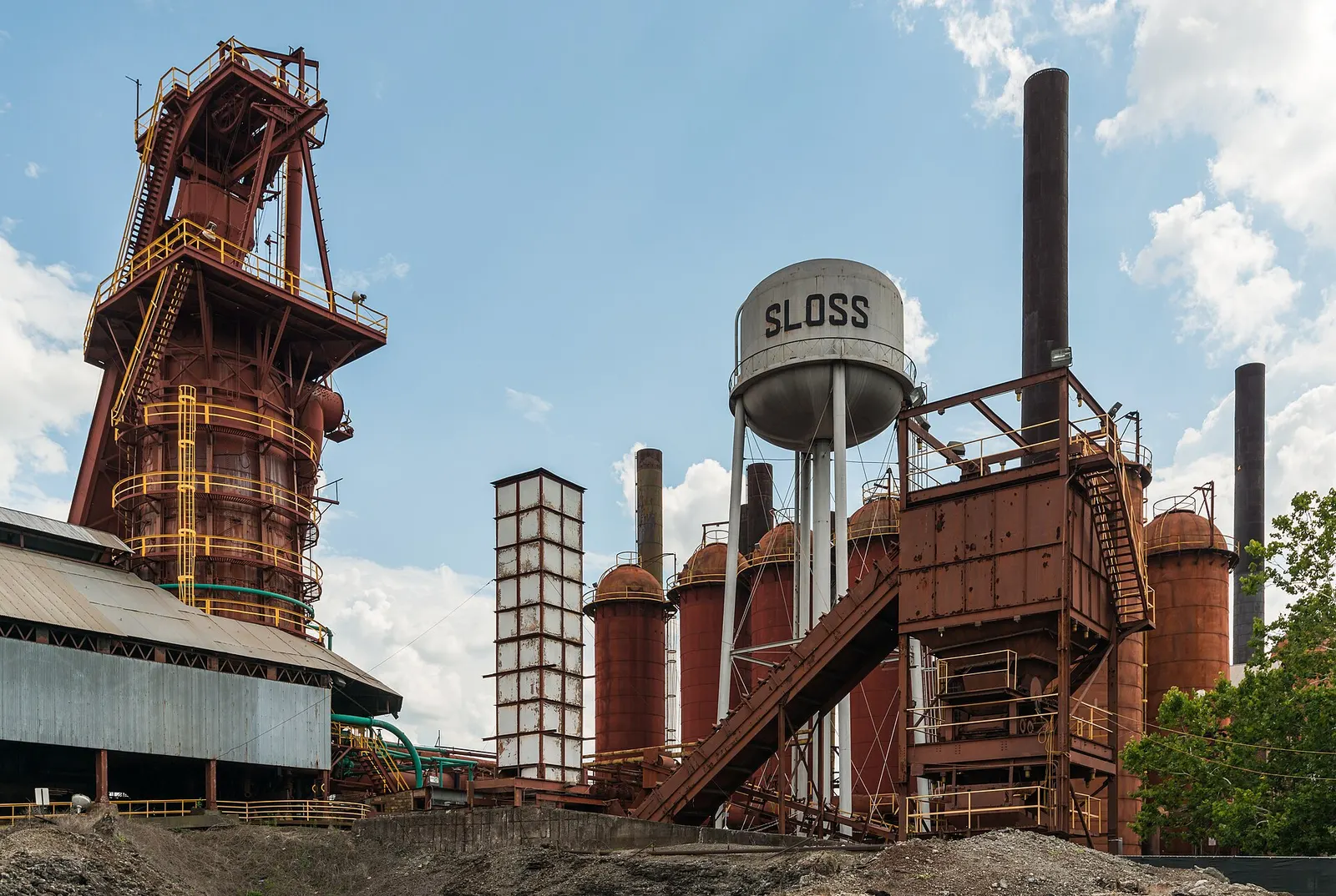 Rust-red blast furnace tower, stoves, smokestacks and a water tower marked SLOSS at the Sloss Furnaces industrial site in Birmingham, Alabama, under a partly cloudy sky