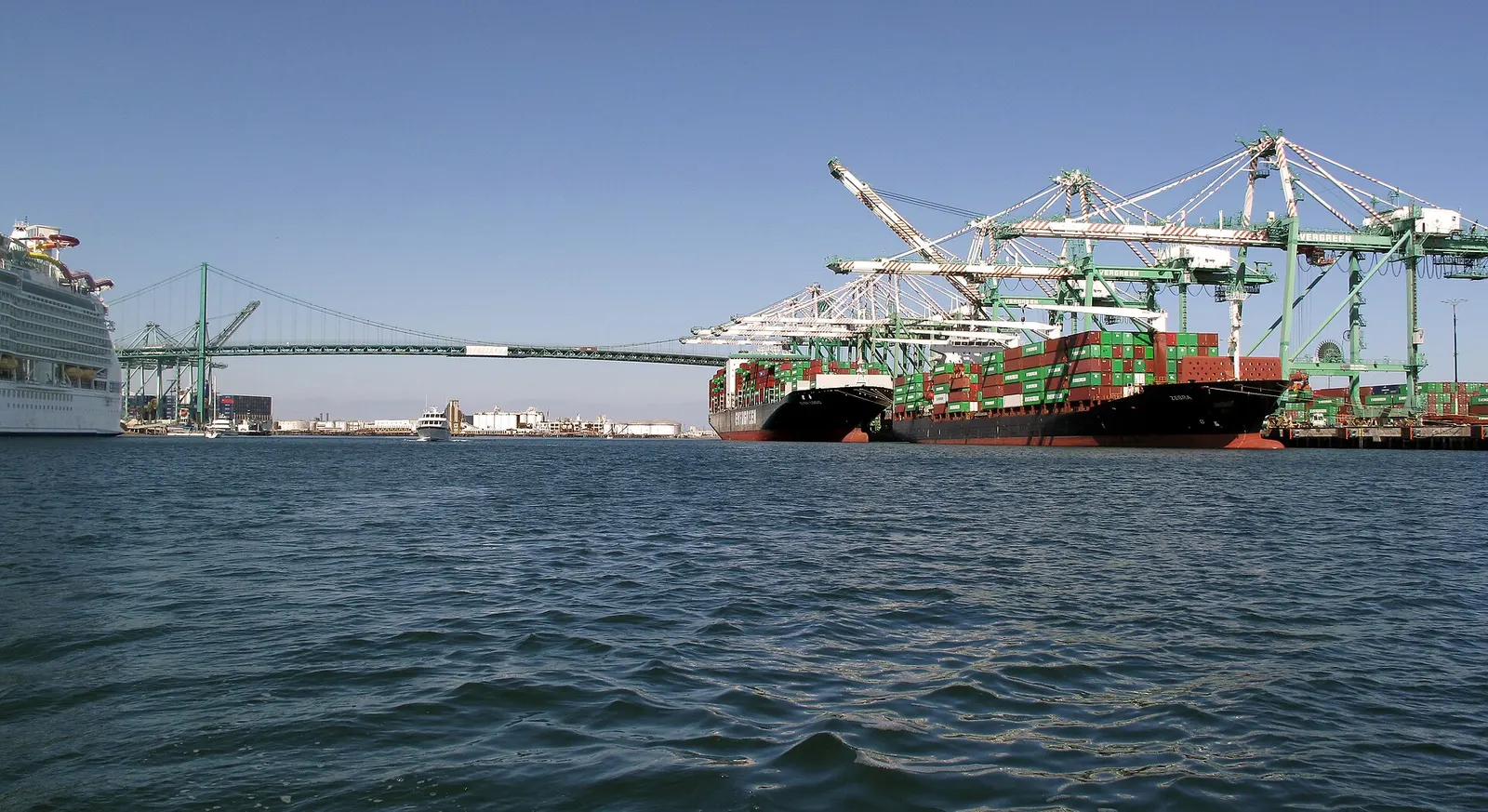 Two loaded container ships beside tall green cranes at the Port of Los Angeles, with a cruise ship at left and a suspension bridge across the harbor under a clear sky