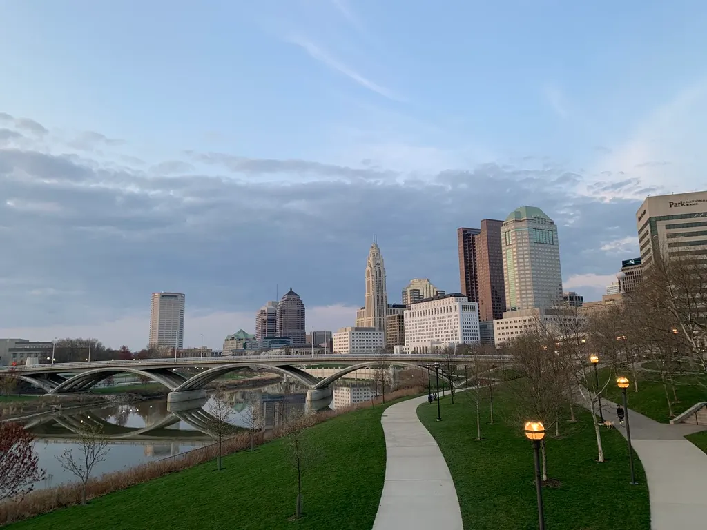 Downtown Columbus skyline at dusk beyond the Scioto River, with an arched bridge over the water and a lamp-lit riverside path in the foreground