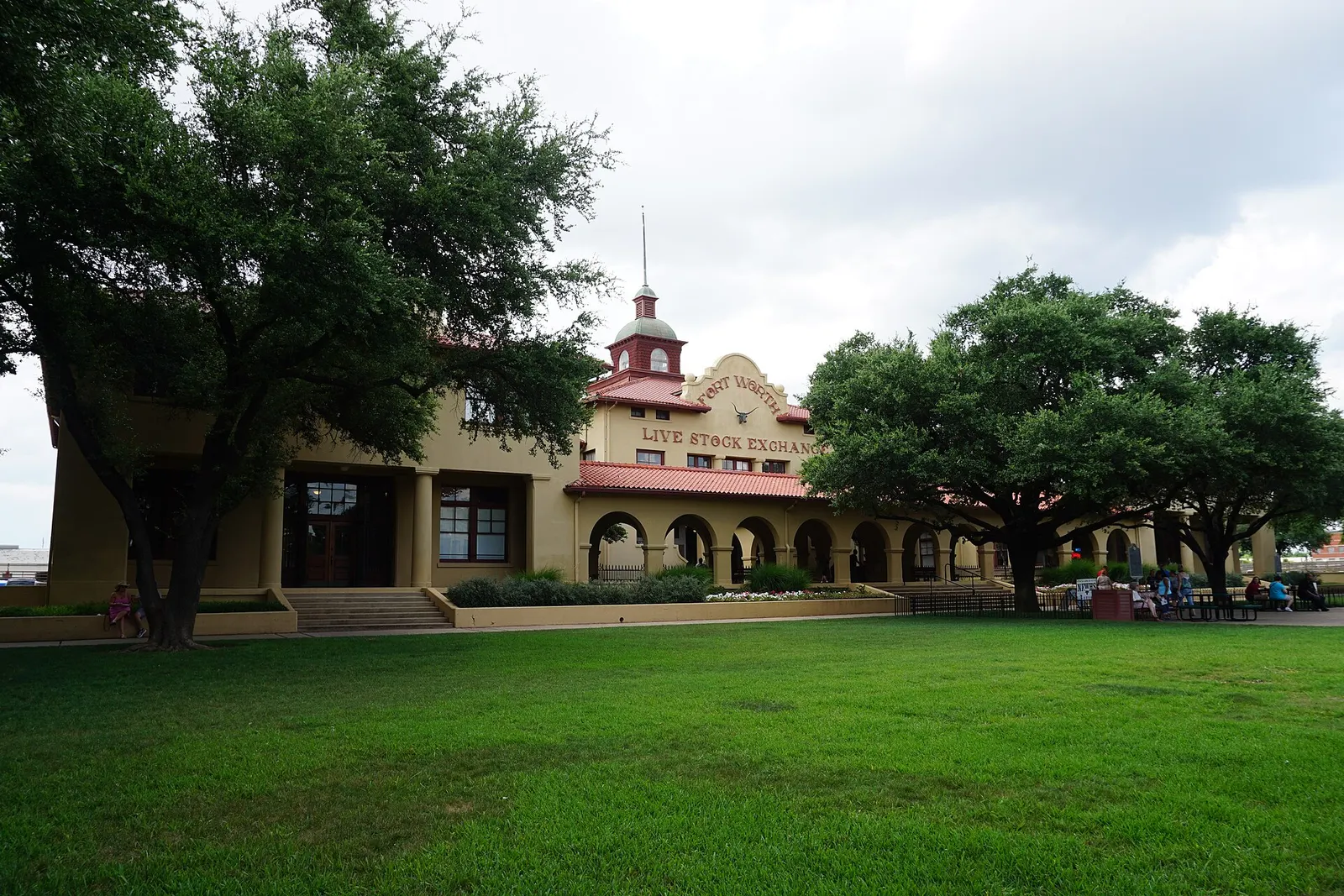 The Livestock Exchange Building in the Fort Worth Stockyards, a tan building with a red tile roof and lettering reading Fort Worth Live Stock Exchange, beside large green trees and a lawn