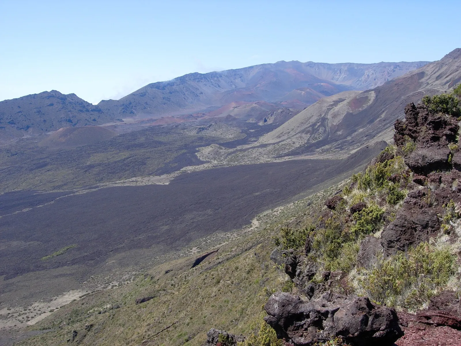 Dark lava flows and red cinder cones across the floor of Haleakala crater on Maui, seen from a rocky rim under a clear blue sky