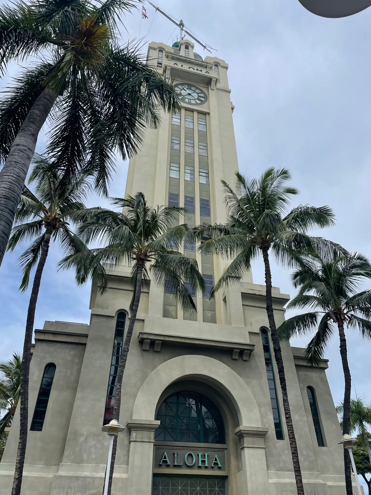 Aloha Tower in Honolulu seen from below: a cream-colored clock tower with the word ALOHA over its arched entrance, framed by tall palm trees under a cloudy sky