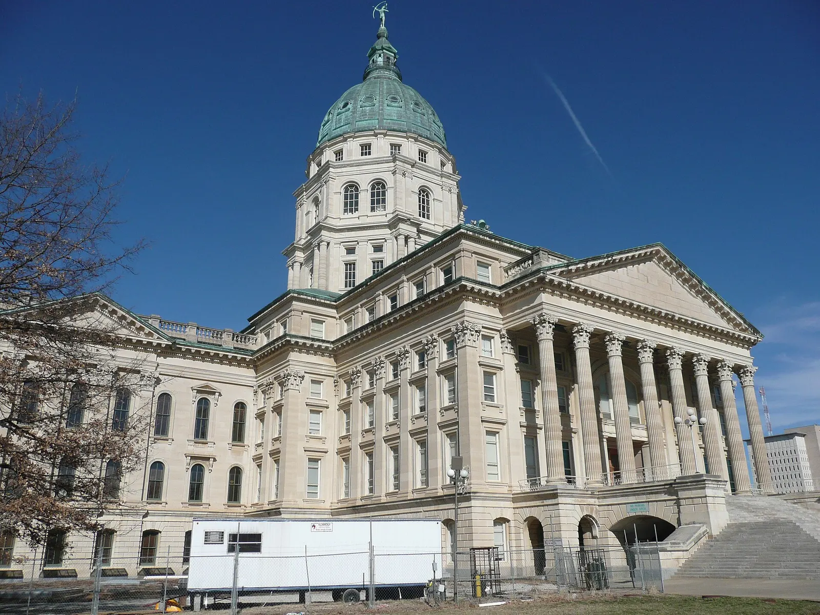 A limestone-colored capitol building with a green copper dome and a front of tall columns, photographed from a corner under a clear blue sky