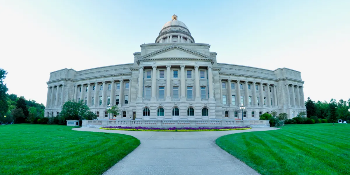The south facade of the Kentucky State Capitol in Frankfort under a pale sky, with a curved walkway, flower bed and mown lawns in front