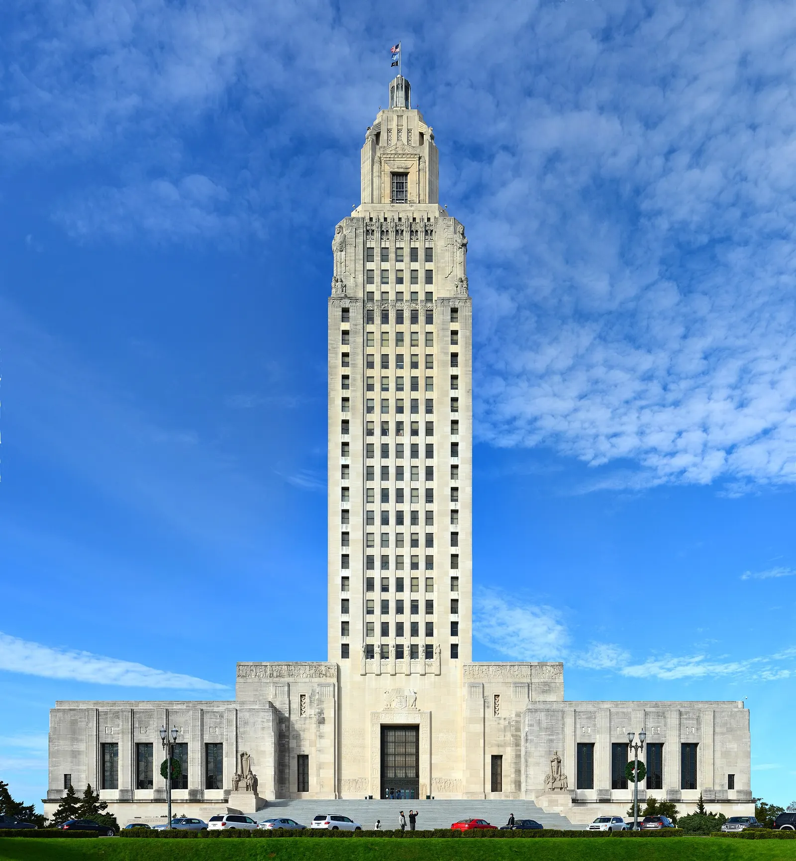 The tall limestone Louisiana State Capitol tower in Baton Rouge rising above a low base, seen from the front lawn under a blue sky with scattered clouds
