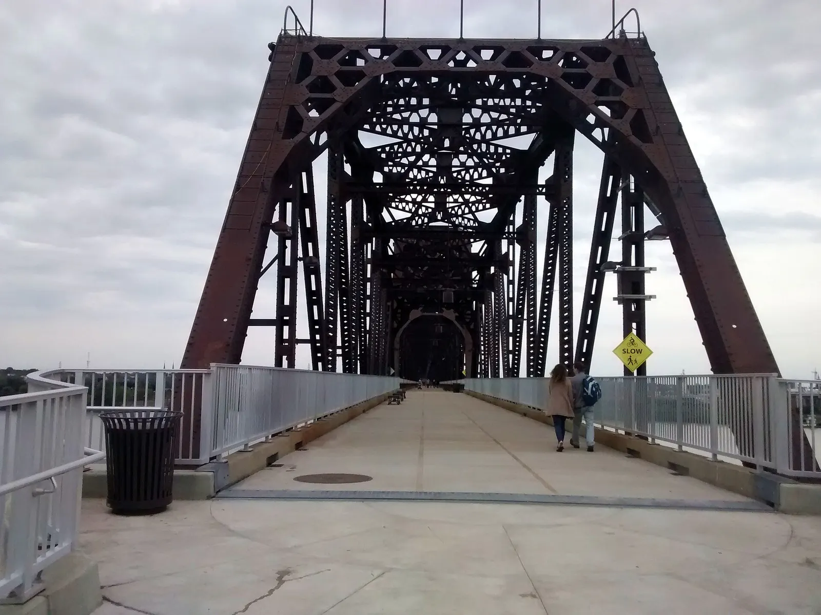A paved pedestrian walkway running into the rust-colored steel truss of the Big Four Bridge in Louisville under an overcast sky, with two people walking away from the camera