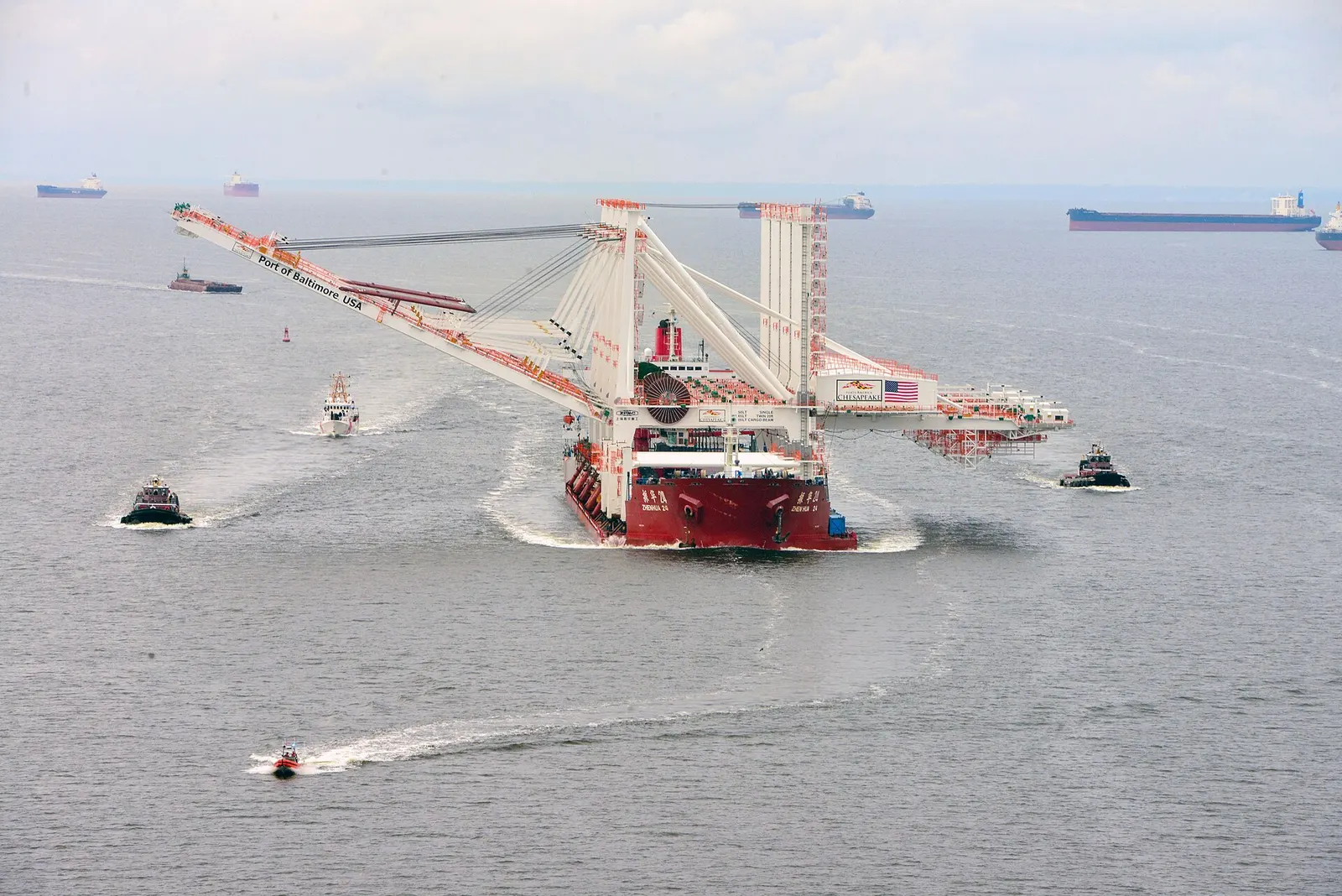 A heavy-lift ship carrying large container cranes marked Port of Baltimore, USA, escorted by tugboats and a Coast Guard boat on the Chesapeake Bay