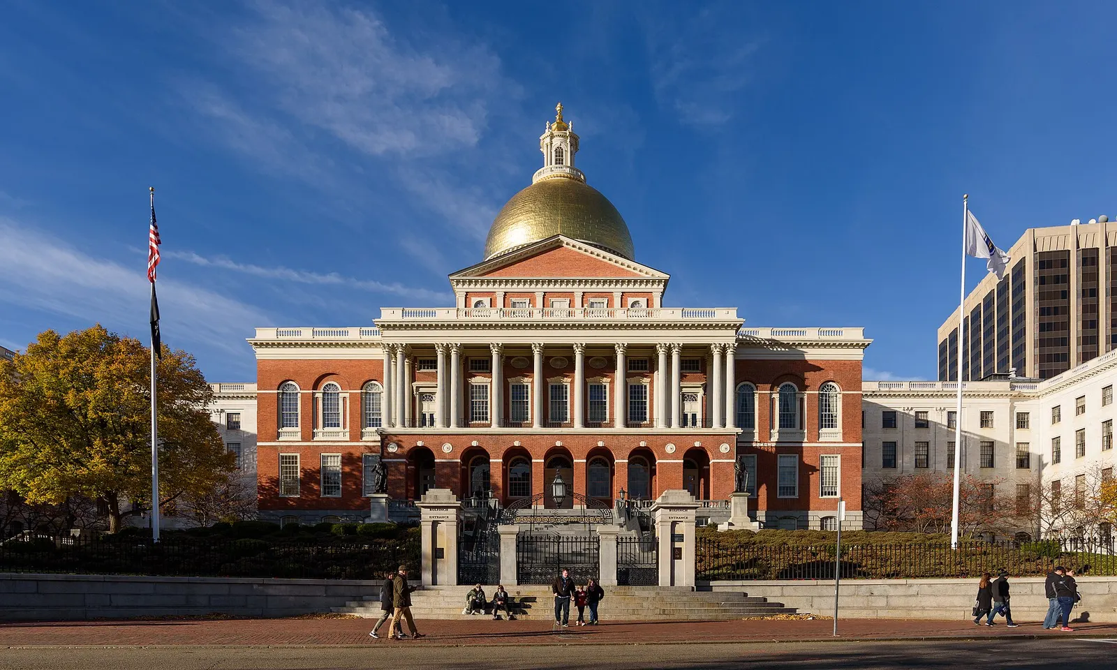 The red-brick Massachusetts State House in Boston with its gold dome and columned portico, seen from across the street on a sunny afternoon with a few pedestrians on the steps