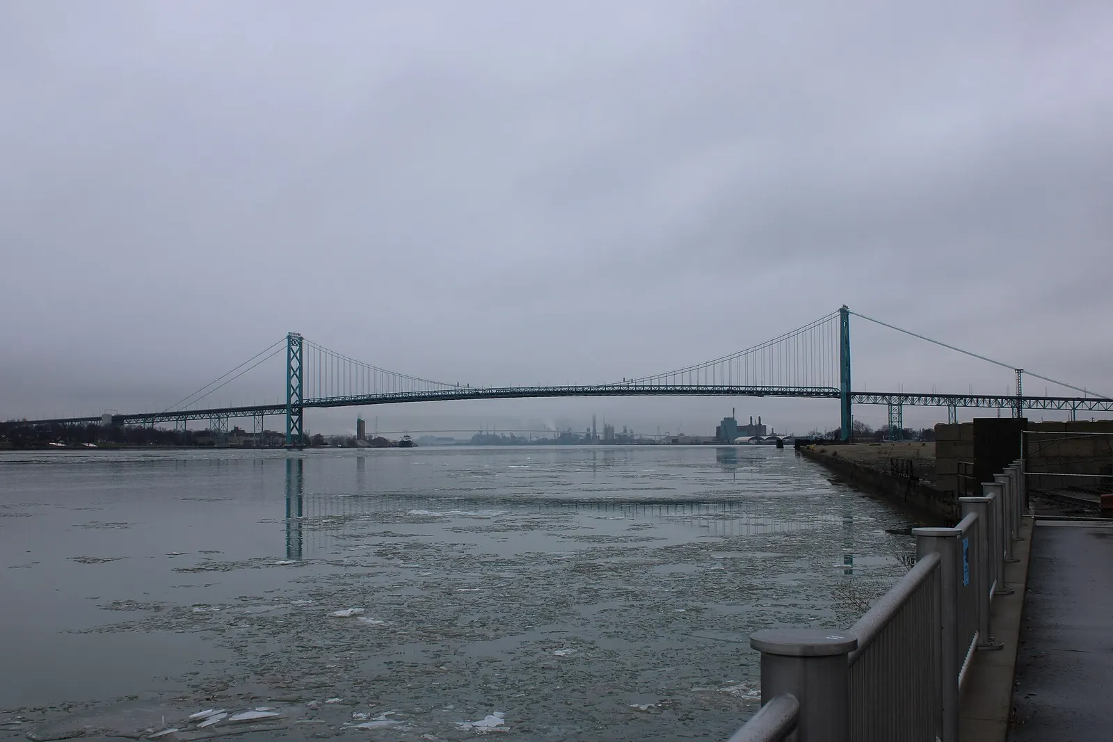 The Ambassador Bridge spanning the Detroit River on a gray winter day, with floating ice on the water and a railing in the foreground