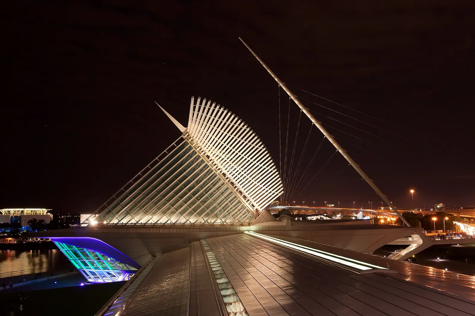 A white winged glass and steel building lit at night beside a harbor, with a tall angled mast and highway lights behind it
