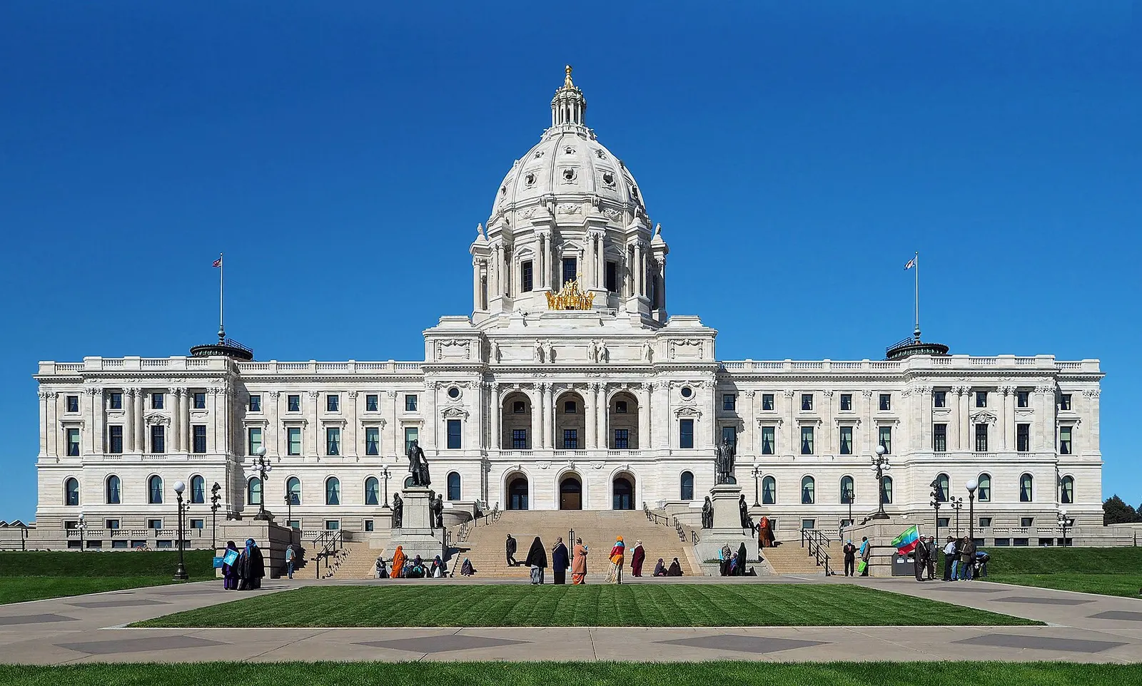Front of the white marble Minnesota State Capitol in St. Paul, with its central dome and a lawn in the foreground under a clear blue sky