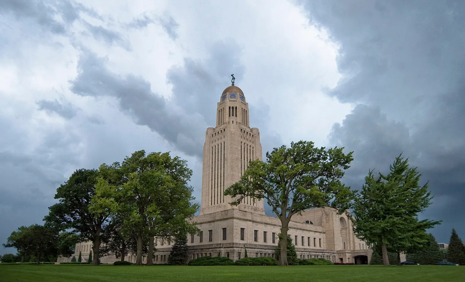 A tall limestone tower topped by a gold dome and a small statue rising behind green trees, under heavy gray storm clouds.