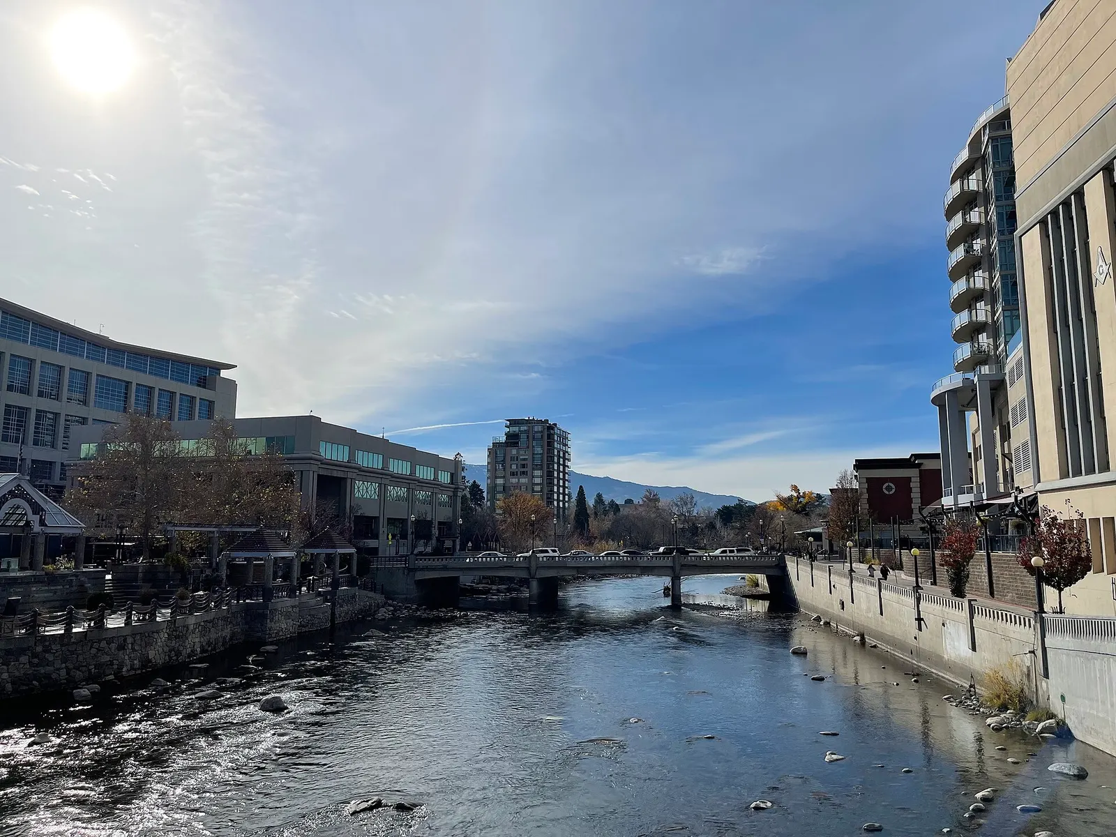 The Truckee River running between downtown buildings in Reno, Nevada, with a low bridge crossing it and mountains in the distance under a blue sky