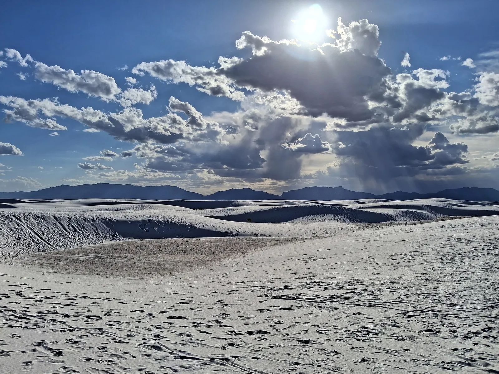 White gypsum sand dunes with footprints under a sky of large clouds and low mountains on the horizon