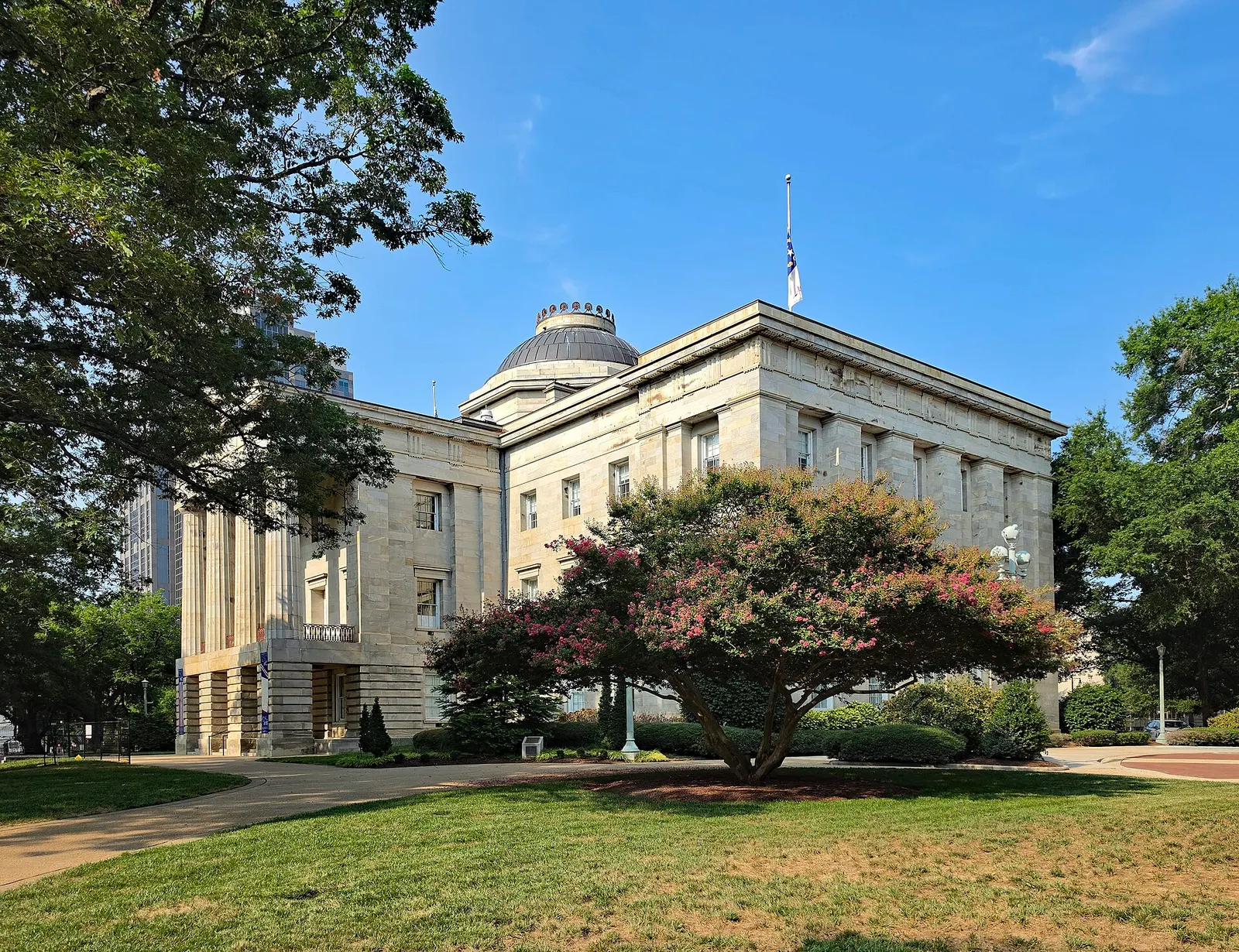 The North Carolina State Capitol in Raleigh, a pale stone Greek Revival building with a low dome, seen from the lawn past a flowering crape myrtle tree under a blue sky