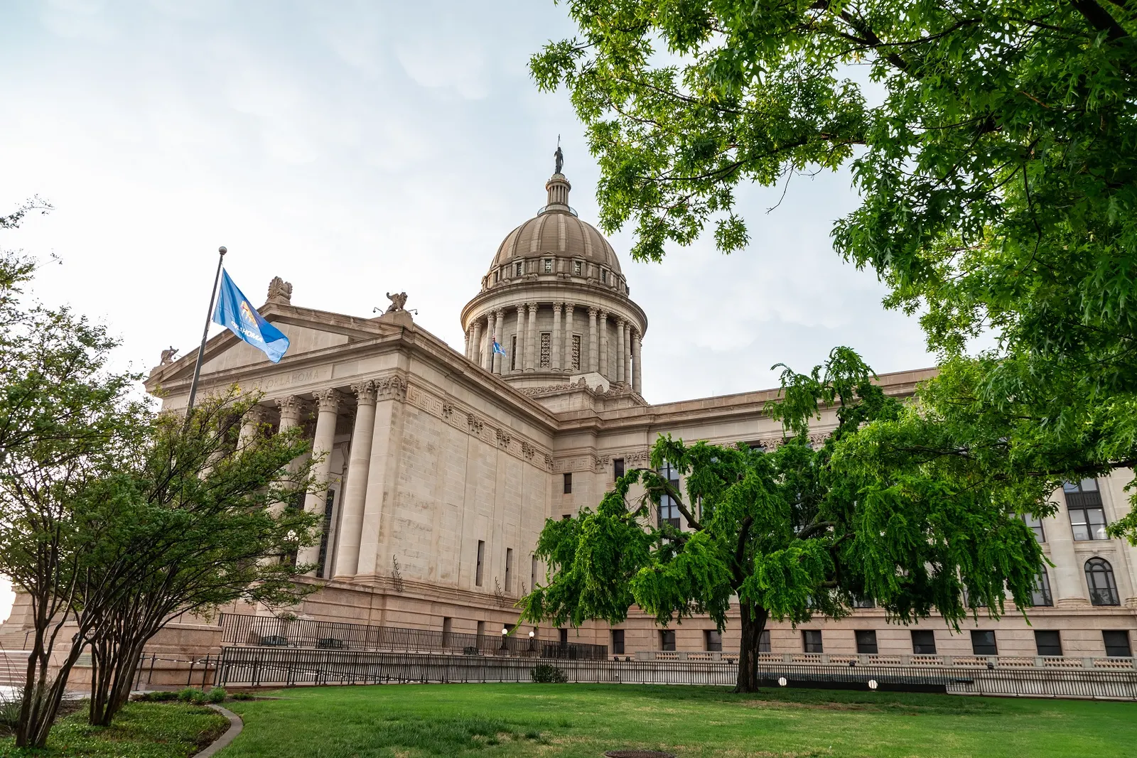 A stone capitol building with a domed tower and tall columns, a blue flag on a pole at the left and green trees in the foreground.