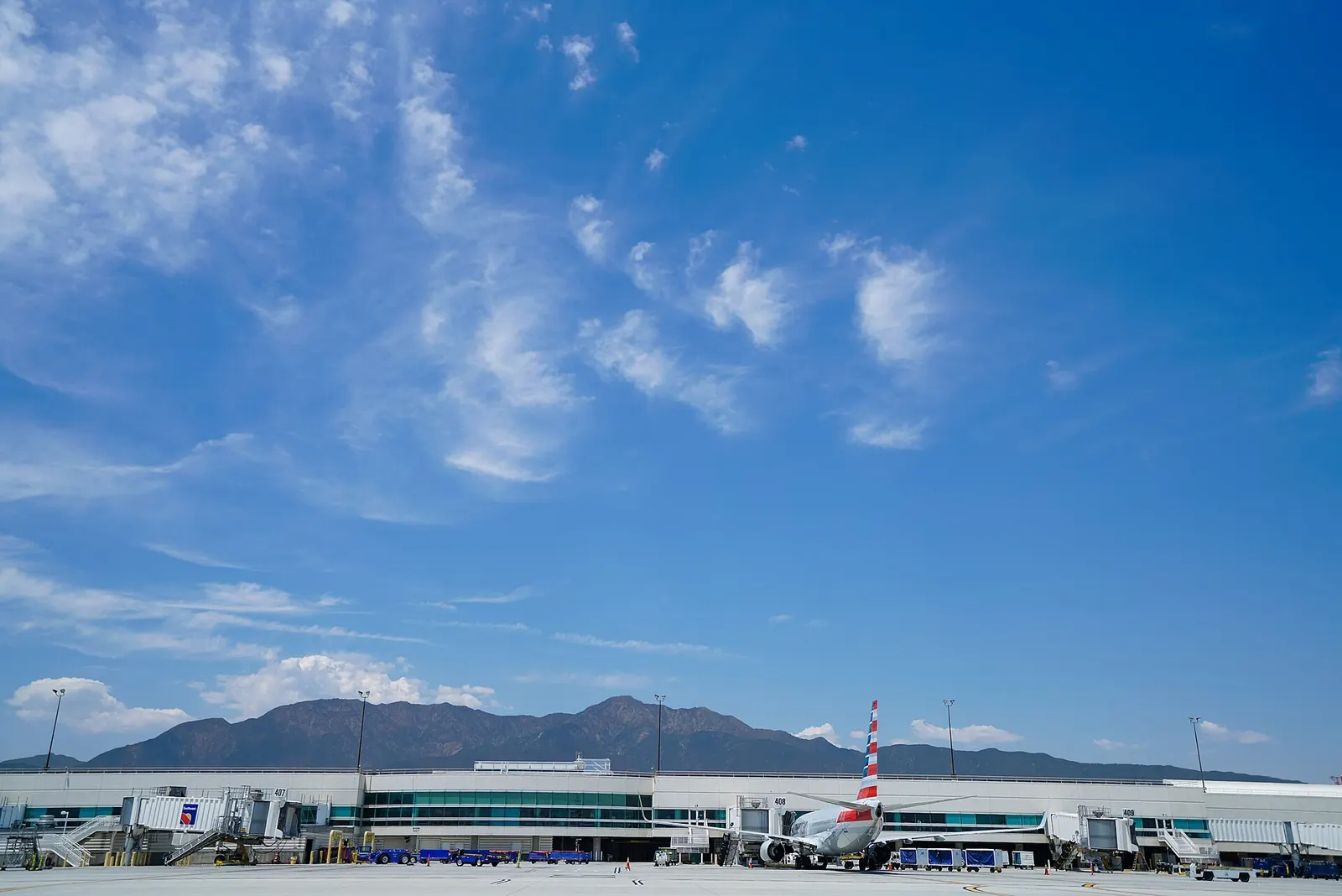 The Ontario International Airport terminal seen from the airfield, an airliner at a gate and the San Gabriel Mountains behind it under a wide blue sky