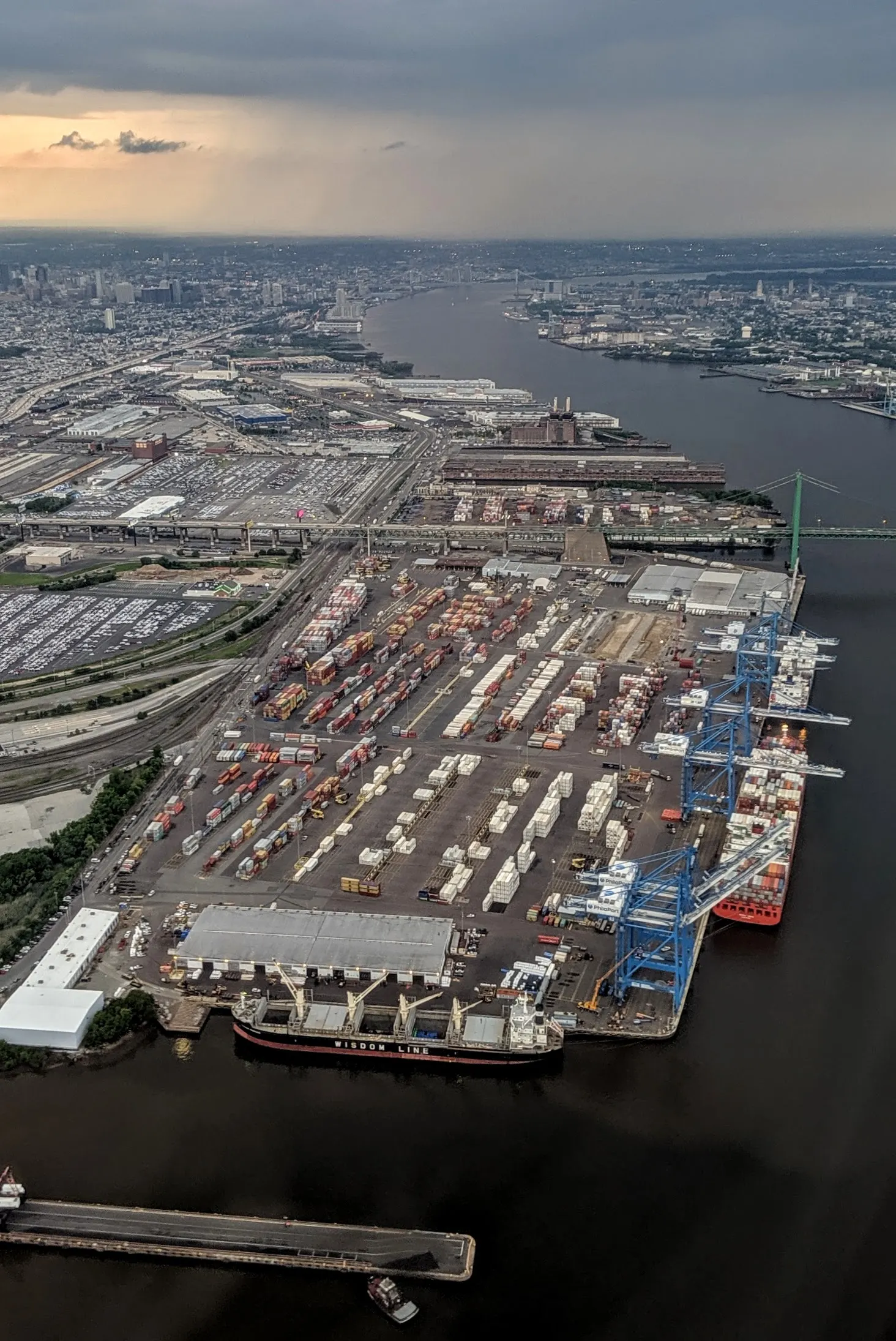Aerial view of the Packer Avenue Marine Terminal in Philadelphia, with container stacks, blue cranes at the berth and a green-towered bridge crossing the Delaware River toward the city