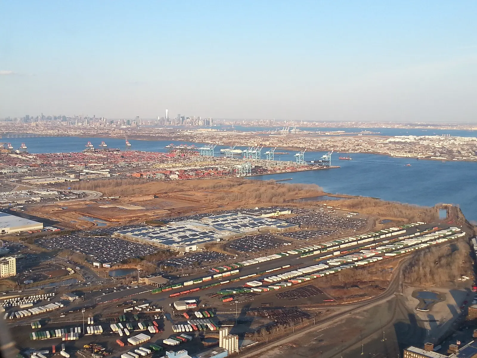 Aerial view over the Port Newark-Elizabeth container terminal, with container yards and blue cranes along the water and the Manhattan skyline faint on the horizon