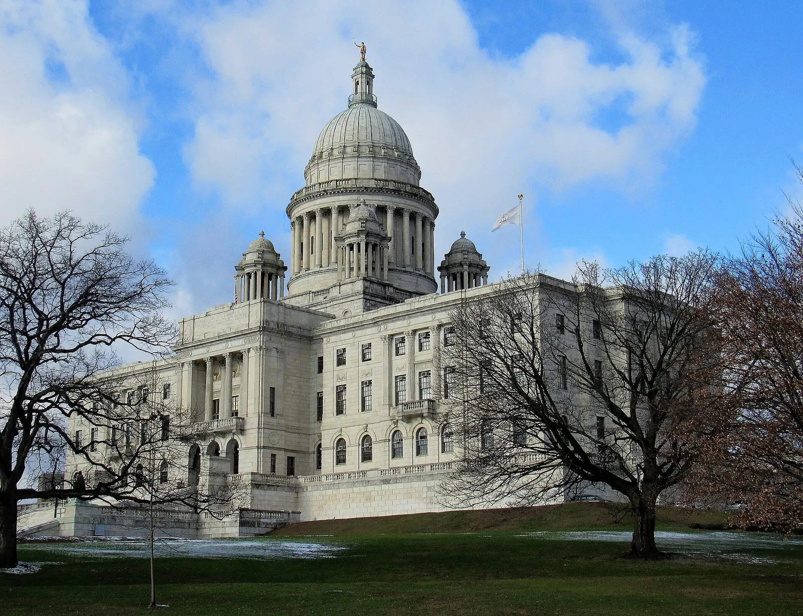 The white marble Rhode Island State House and its dome rising over bare trees and a lawn under a partly cloudy sky