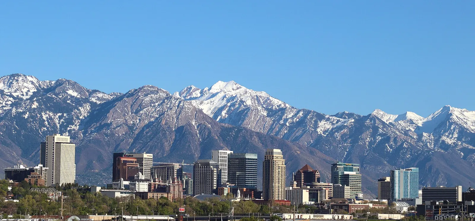 Downtown Salt Lake City towers in front of the snow-capped Wasatch Range under a clear blue sky.