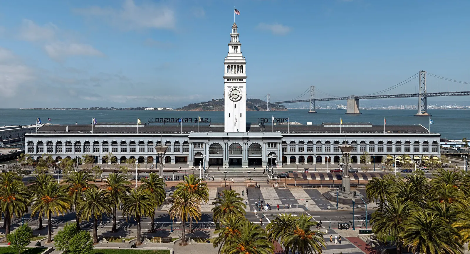 The San Francisco Ferry Building and its white clock tower along the Embarcadero, with palm trees in the foreground and the Bay Bridge crossing the bay behind
