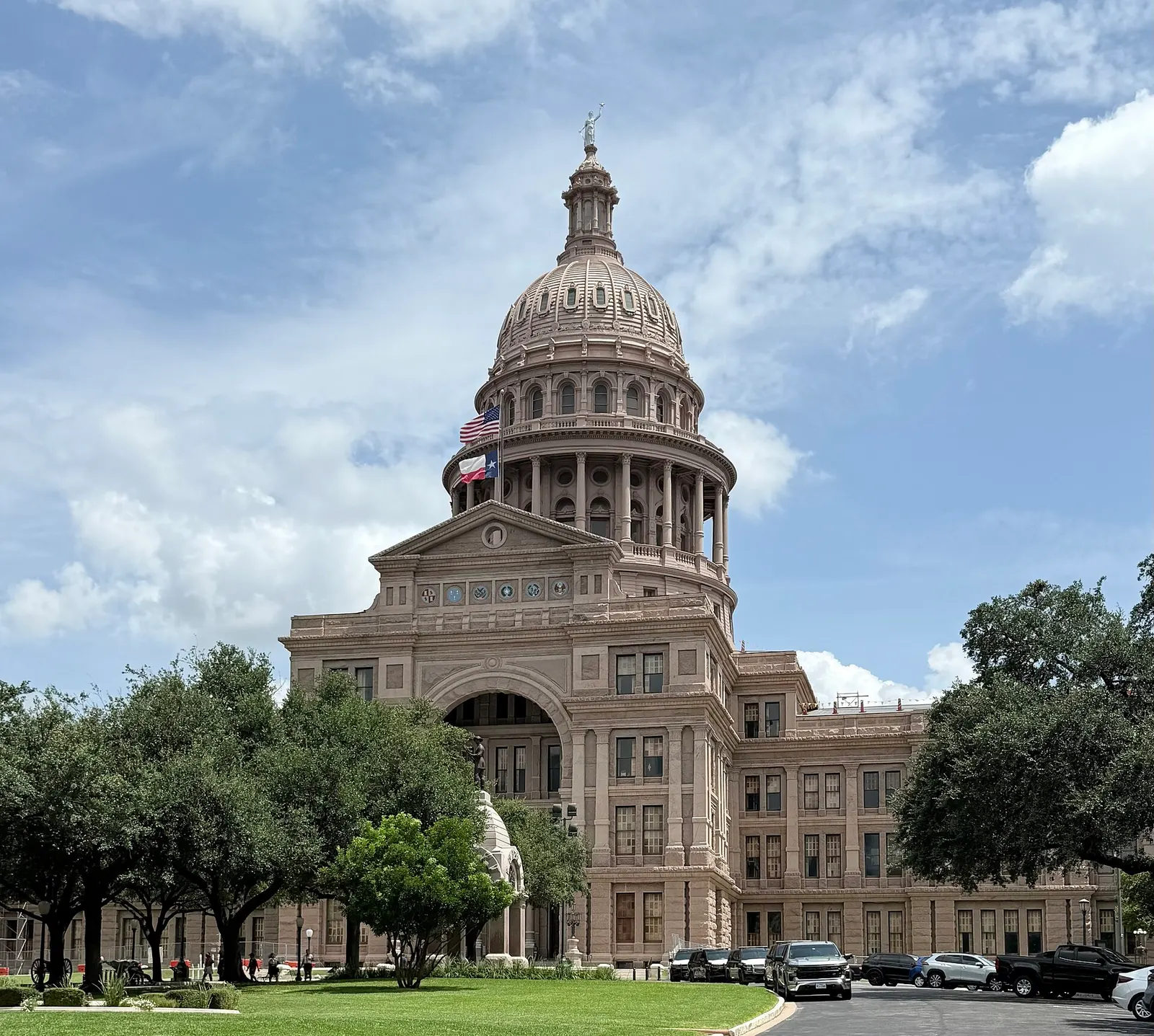 The domed Texas State Capitol in Austin behind a lawn and live oak trees, under a partly cloudy sky