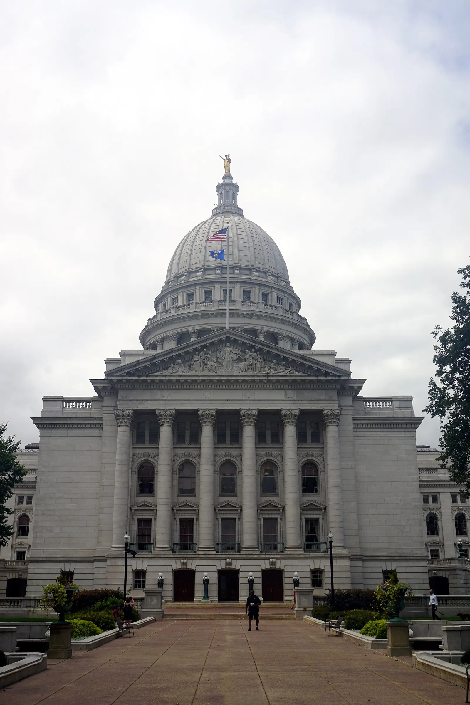 The Wisconsin State Capitol in Madison seen from the front, with its white granite dome, columned facade and a flag flying under a cloudy sky.